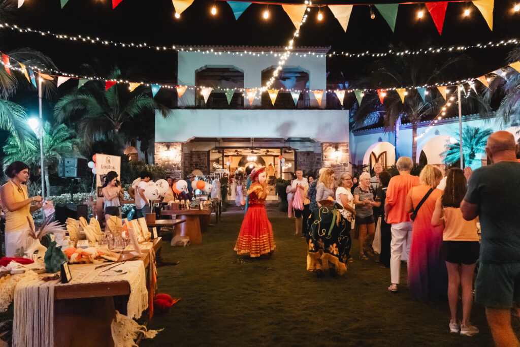 A lively Mercadito outdoor night market with people browsing stalls, colorful bunting and string lights overhead, palm trees in the background, and a performer in a red and yellow dress celebrating local culture.