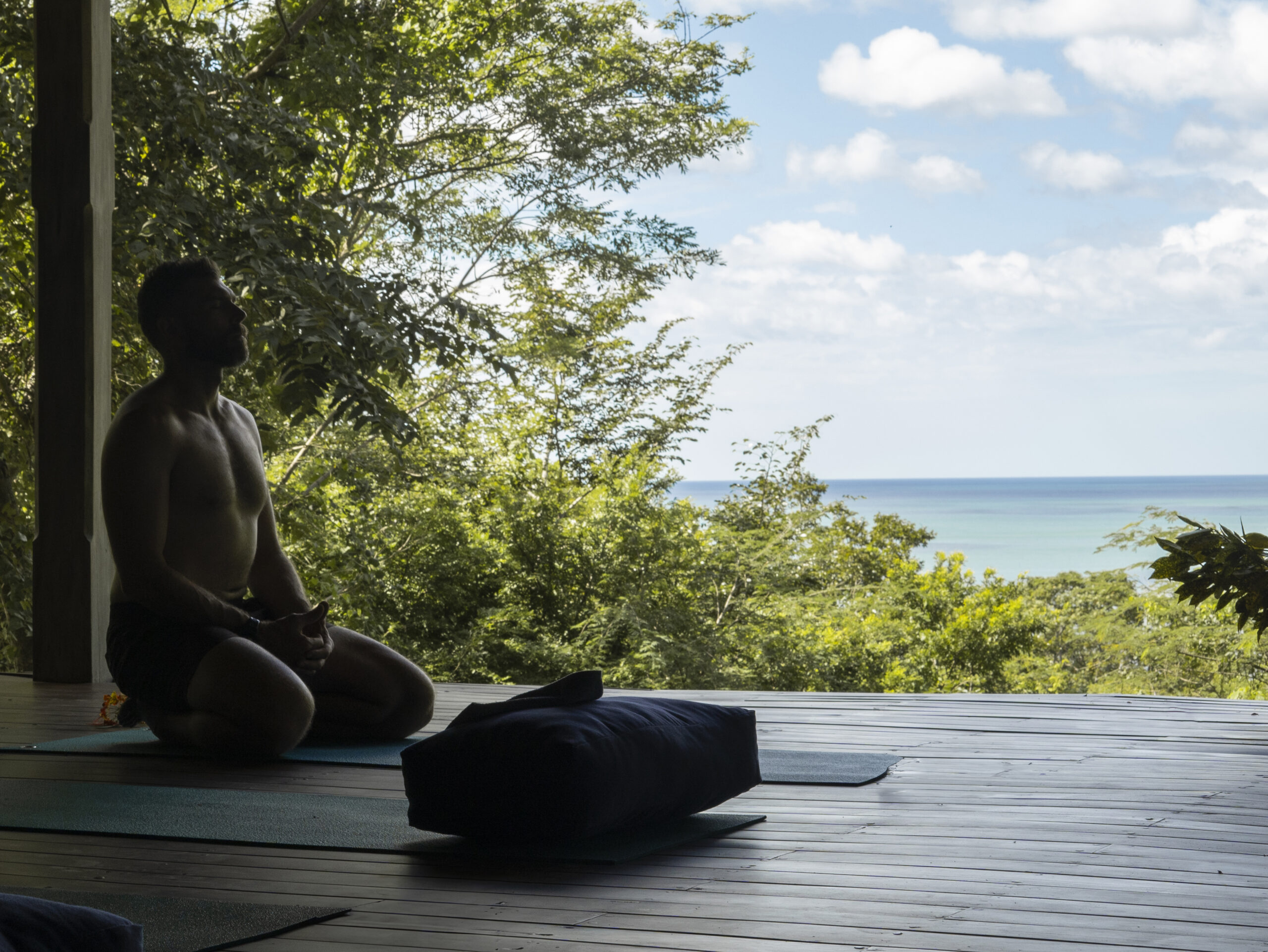A person sits cross-legged on a yoga mat in a shaded wooden area, practicing mindfulness as they face lush greenery and the ocean under a bright, cloudy sky, appearing to meditate or relax quietly.