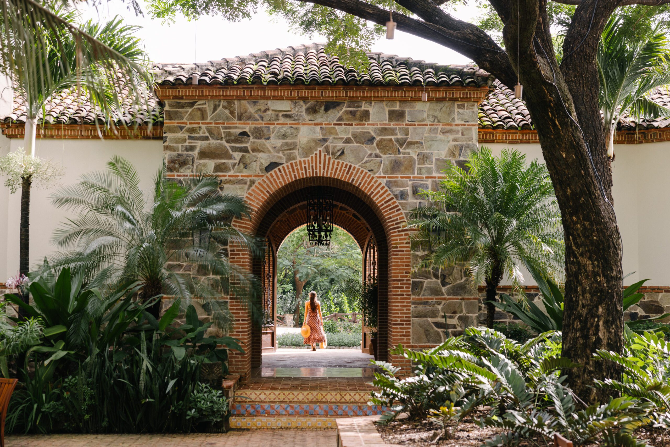 A woman in a yellow dress walks through a stone archway of a building with a tiled roof, surrounded by lush green plants and trees.