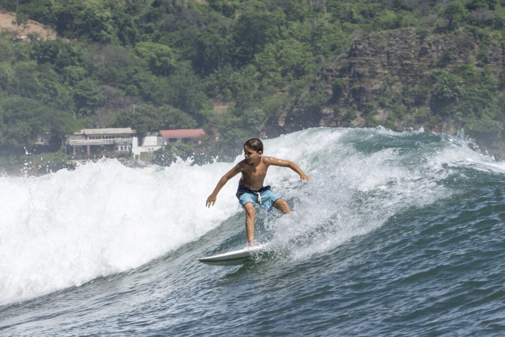A young boy in blue swim trunks is surfing on a large wave near the shore, with green trees and a few buildings visible in the background under a clear sky.