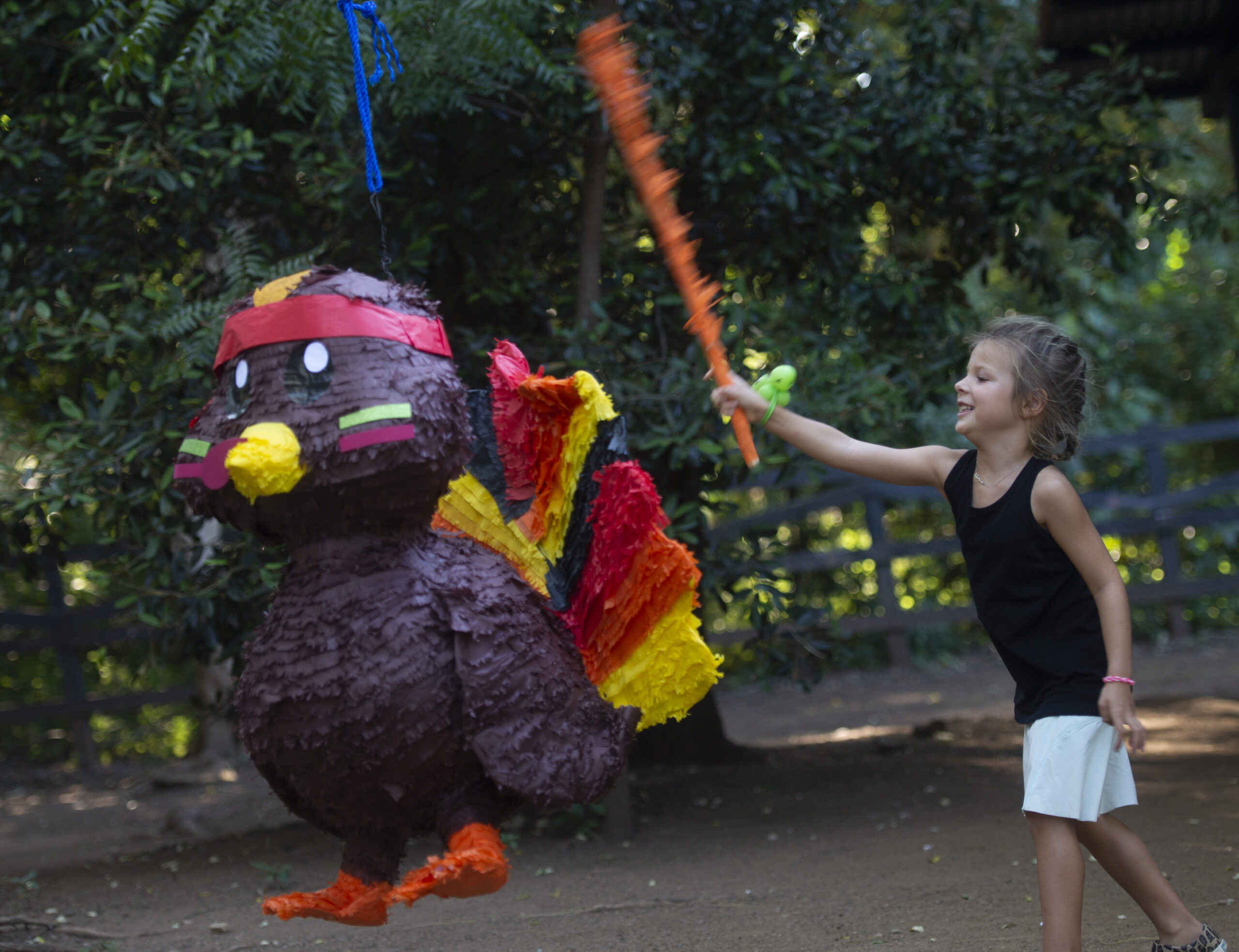 A young girl in a black tank top and white shorts swings a stick at a large turkey-shaped piñata outdoors, surrounded by greenery—its the perfect way to Give Thanks in Paradise.