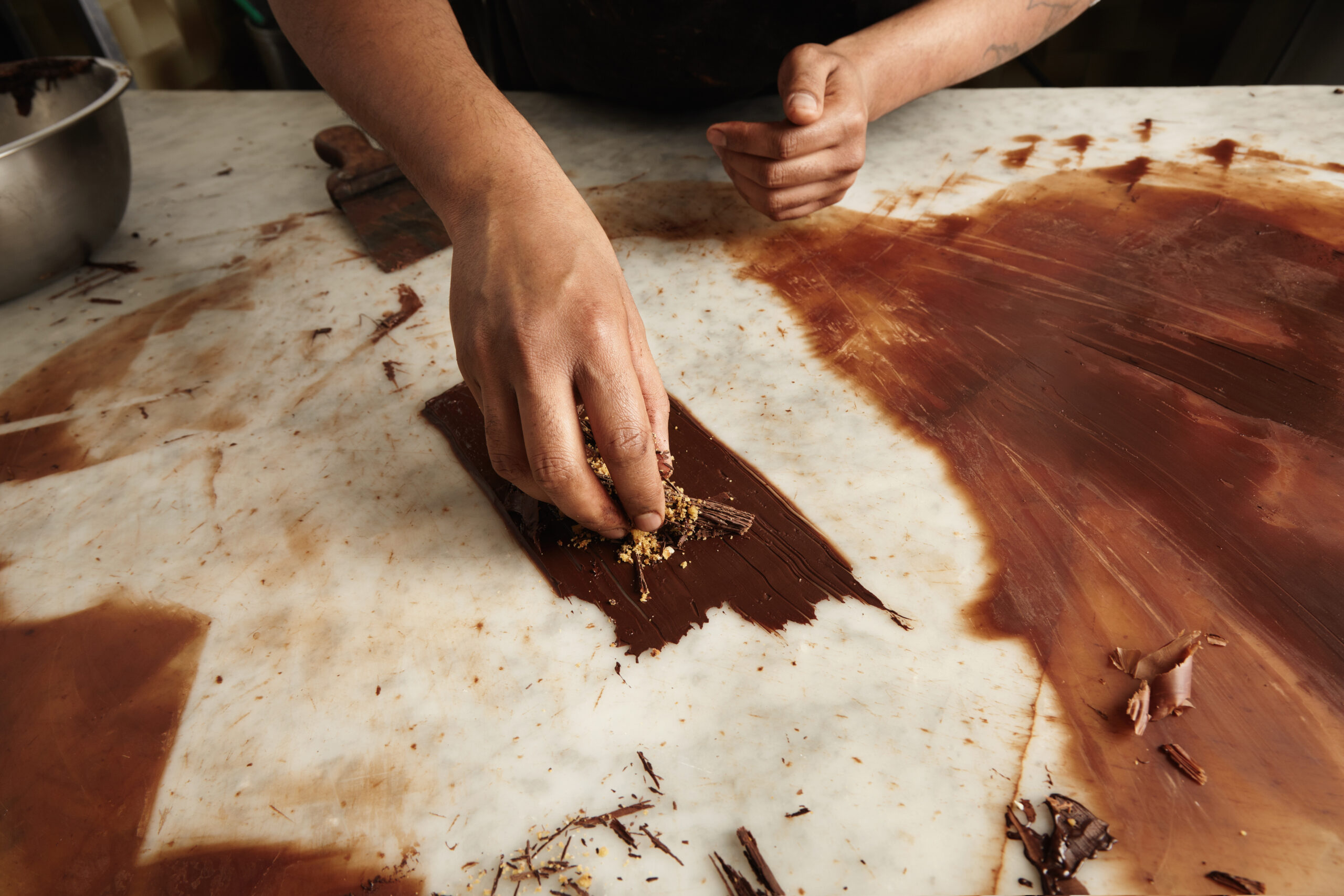 A person’s hand sprinkles chopped nuts or toppings onto a layer of melted chocolate spread over a marble countertop, with chocolate smears and a metal bowl nearby—perfect for creating Thanksgiving treats on the Emerald Coast.