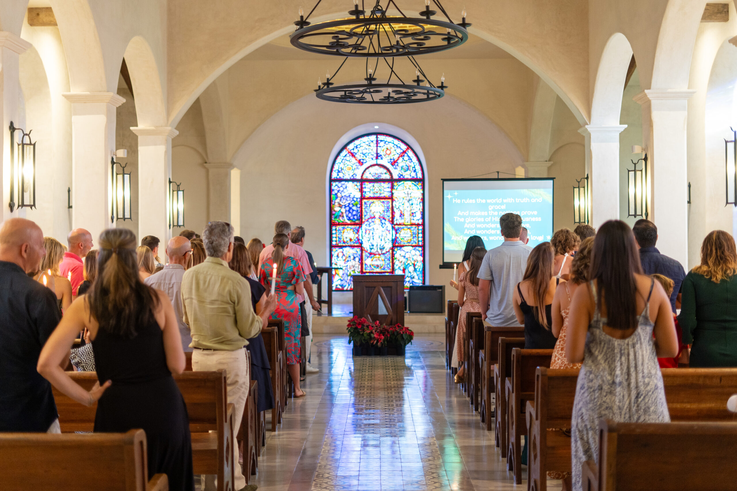 People standing in pews inside a church with arched ceilings on the Emerald Coast, facing a large stained glass window and text on a screen. Warm lighting and candles evoke a reverent Holiday Spirit.