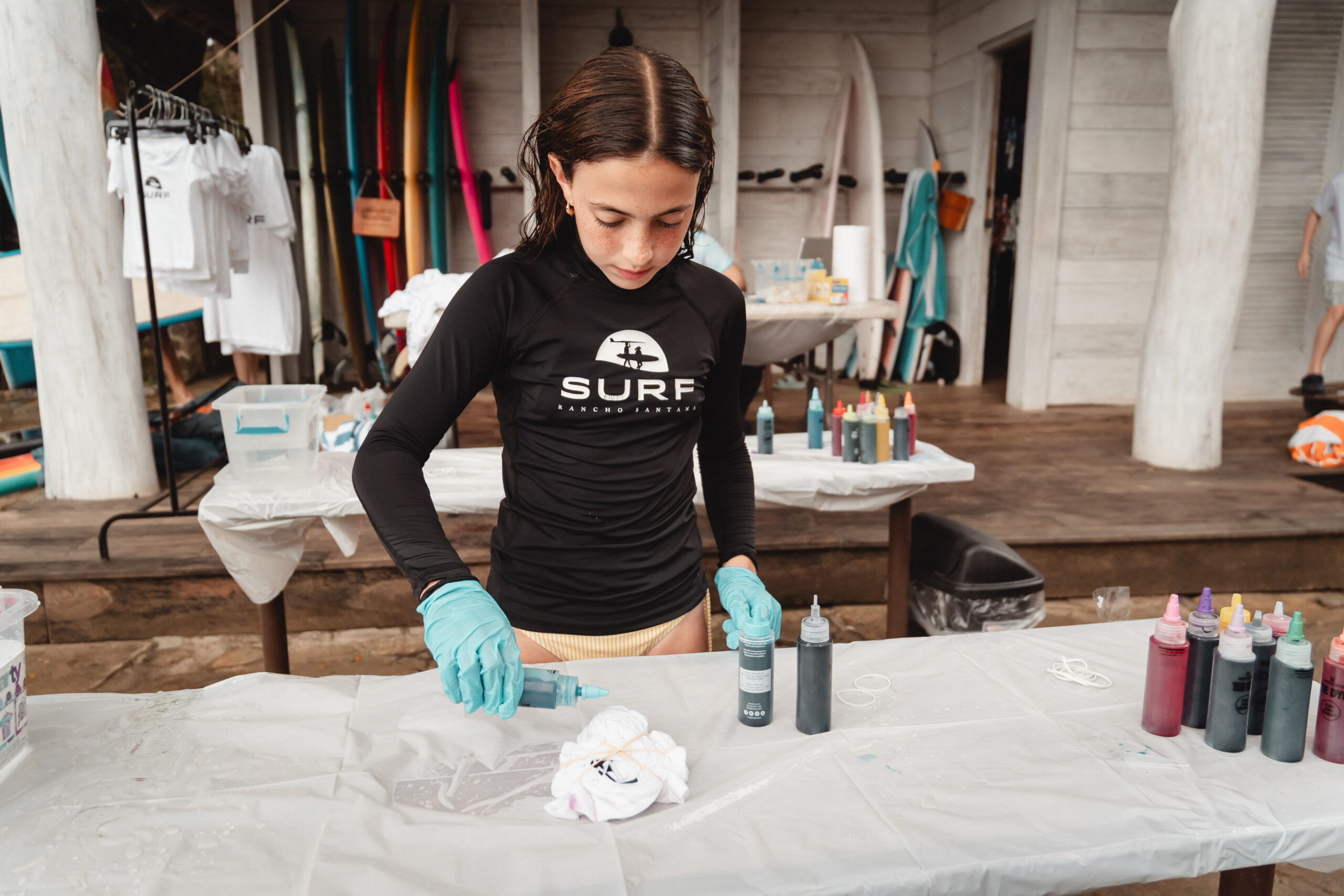 A person wearing gloves and a black SURF shirt applies dye to a shirt on a table covered with plastic, surrounded by bottles of dye and surfboards in the background, crafting timeless memories with coastal elegance.