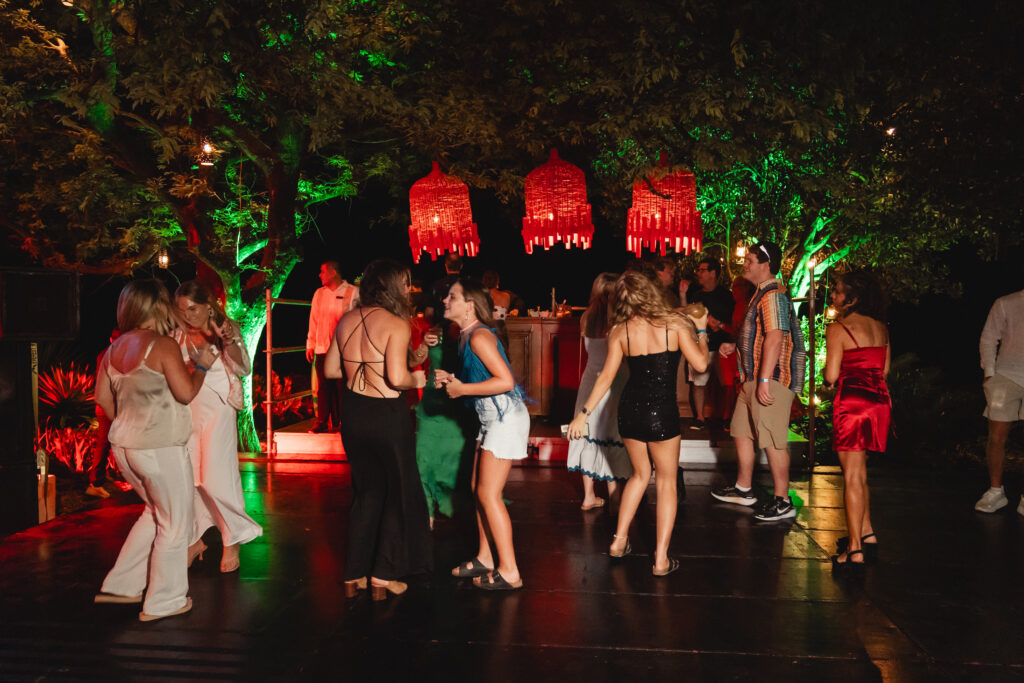 A group of people dance outdoors at night under red lanterns and colorful green and red lights, surrounded by trees. Dressed in semi-formal attire, they celebrate the New Year, creating timeless memories in a scene of coastal elegance.