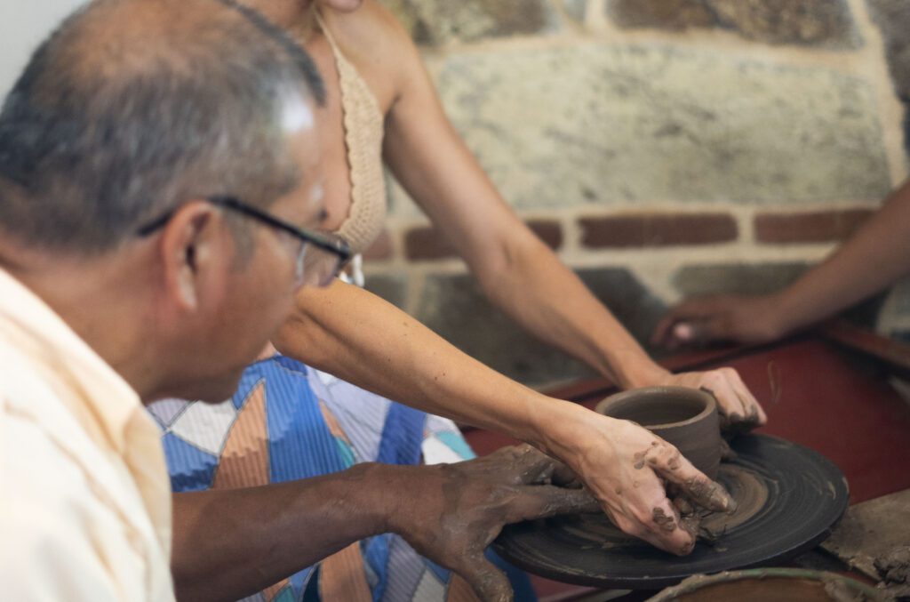 A Chorotega Artisan is guiding another persons hands as they shape clay on a pottery wheel, both hands covered in wet clay. A stone wall is visible in the background, capturing the tradition of creating ceramics.
