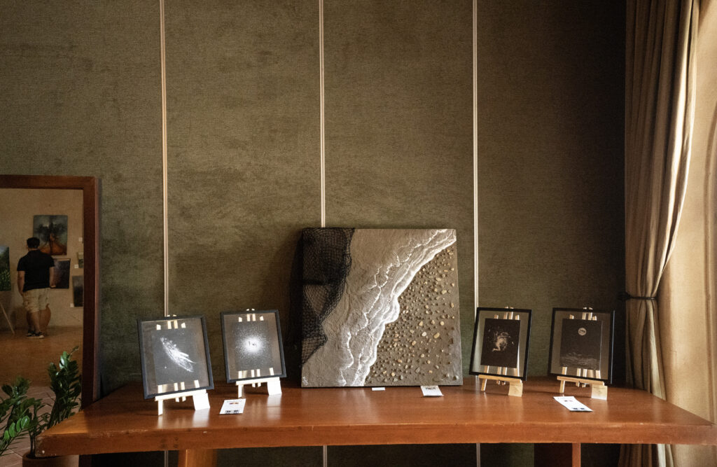 A wooden table displays five framed artworks at a Nicaraguan Art Exhibition, with a central textured painting resembling a shoreline and four smaller black-and-white pieces on easels. A person is reflected in a mirror on the left side of the room.