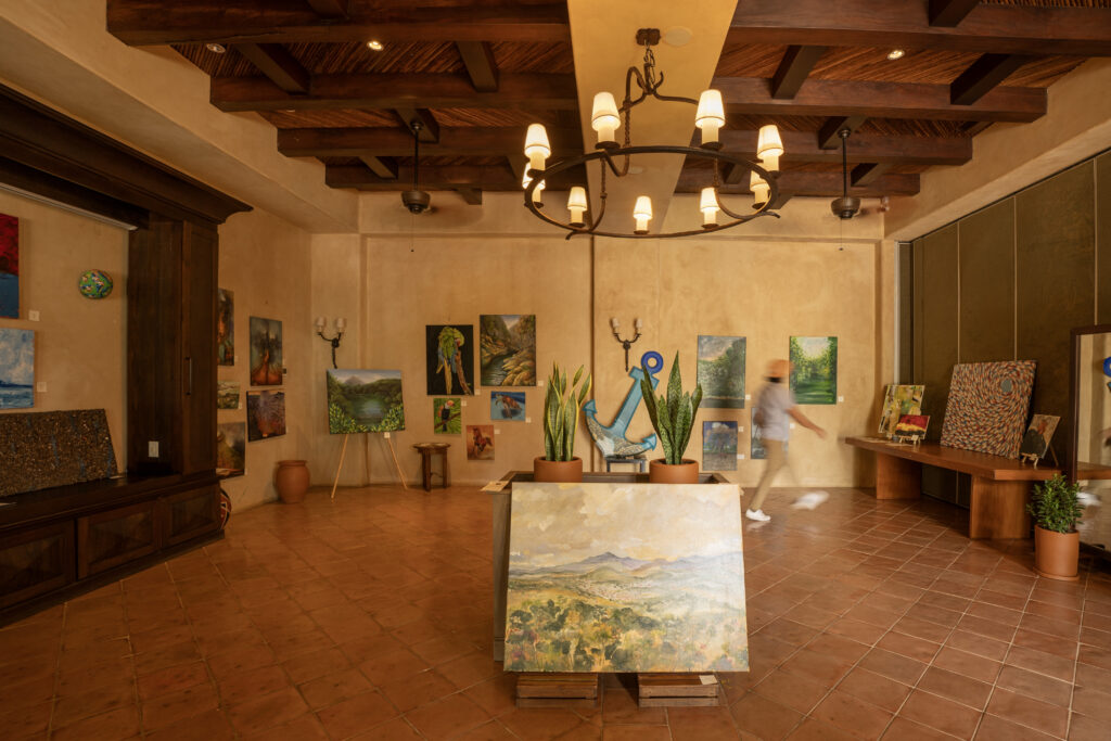 A warmly lit art gallery with tan walls and a tiled floor hosts a Nicaraguan Art exhibition. A person walks past landscape and abstract paintings, while potted plants and a chandelier hanging from the wooden ceiling complete the scene.