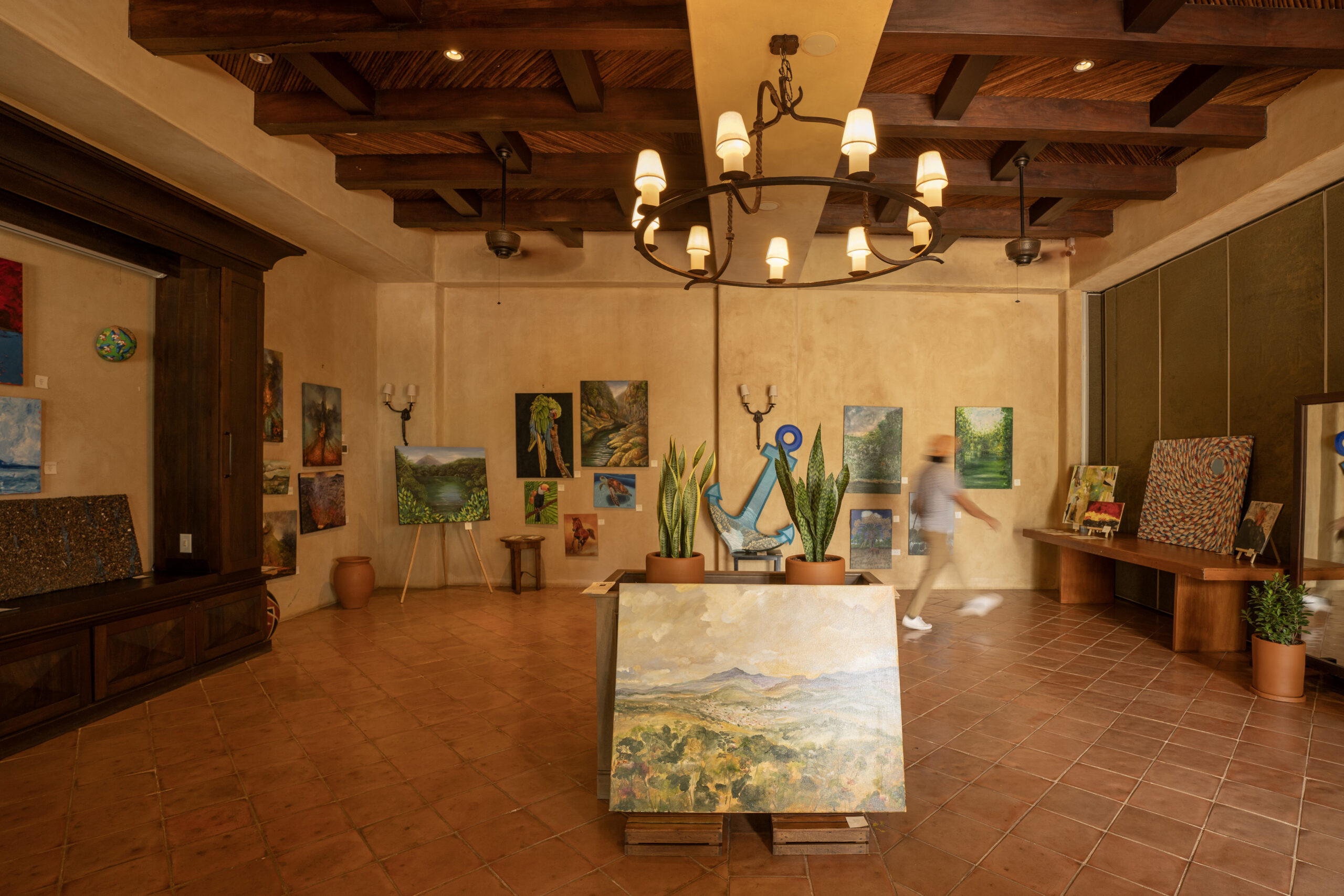 A warmly lit art gallery with tan walls and a tiled floor hosts a Nicaraguan Art exhibition. A person walks past landscape and abstract paintings, while potted plants and a chandelier hanging from the wooden ceiling complete the scene.