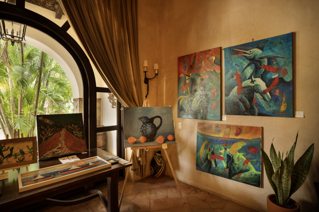 An indoor Nicaraguan Art Exhibition features colorful paintings on beige walls, a potted plant, an easel with a painting of a pitcher and oranges, a wooden table with booklets, and sunlight streaming through a large arched window.