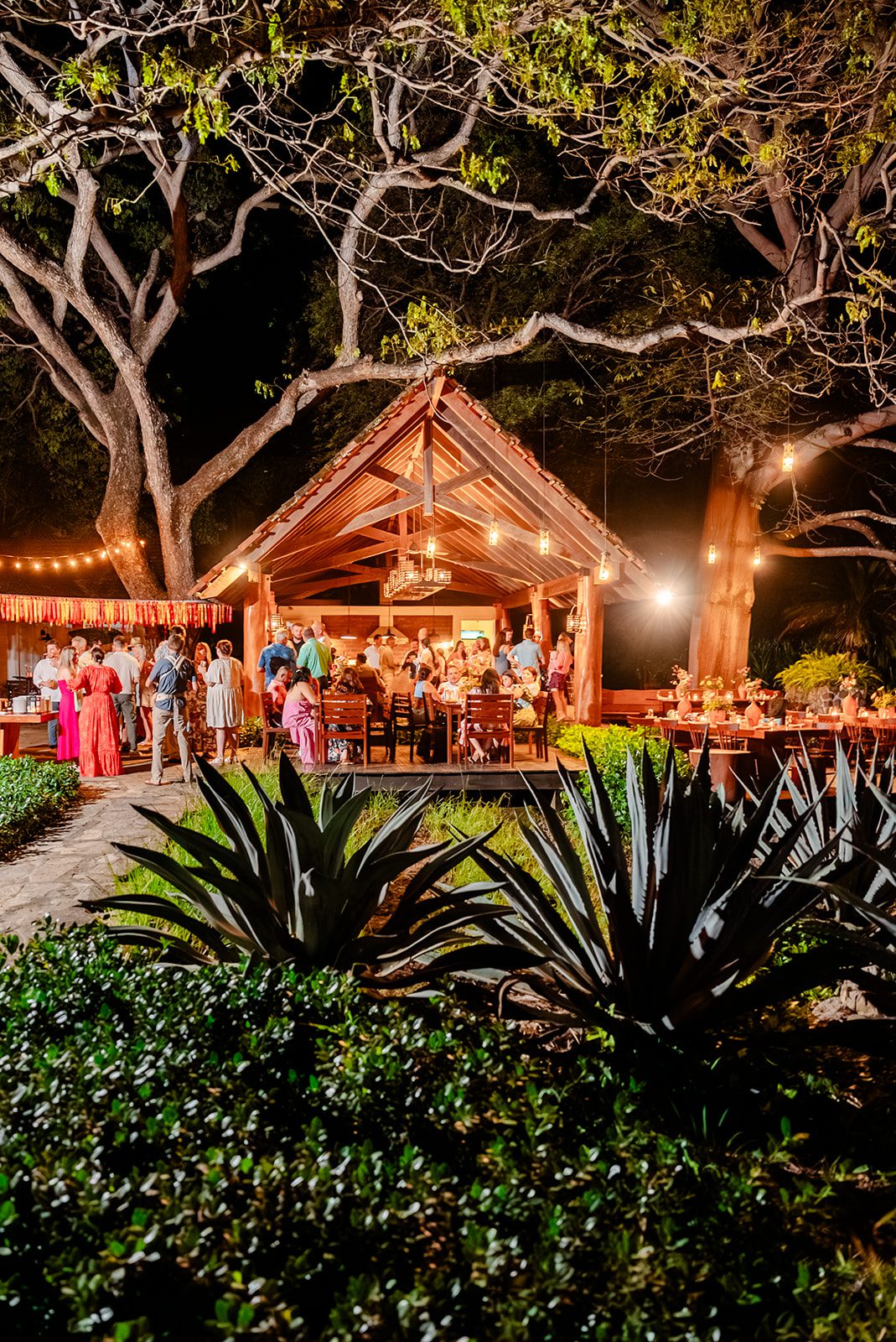 People gather at a warmly lit outdoor pavilion at night, surrounded by large trees and plants. String lights and festive decorations create a lively atmosphere, with guests socializing and dining at tables.
