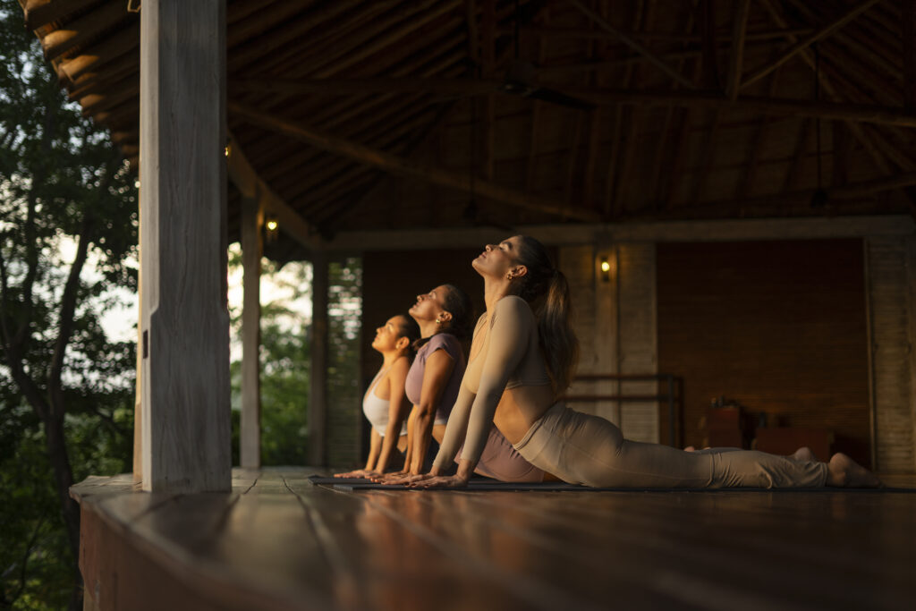 Three women practice yoga and breathwork together on a wooden deck at sunset, performing the upward-facing dog pose. Warm light shines on them, and trees are visible outside the open structure.