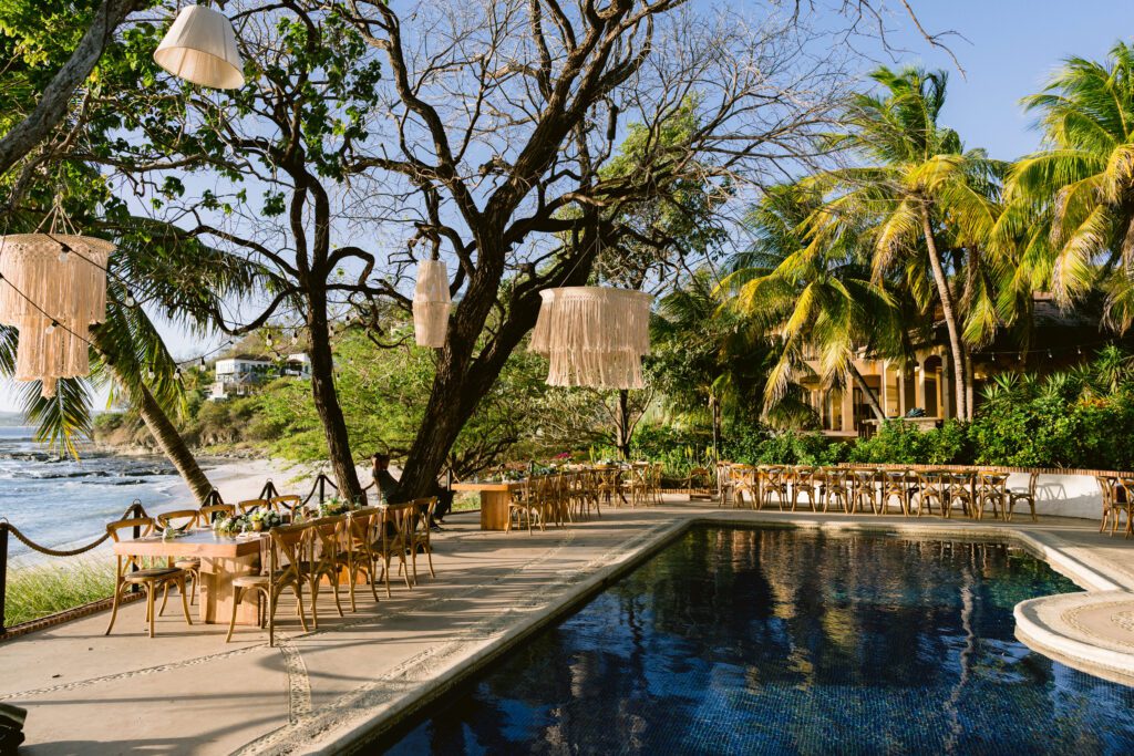 Outdoor dining area beside a pool, with wooden tables and chairs under trees and hanging tasseled lamps, overlooking the ocean and surrounded by lush greenery and palm trees.
