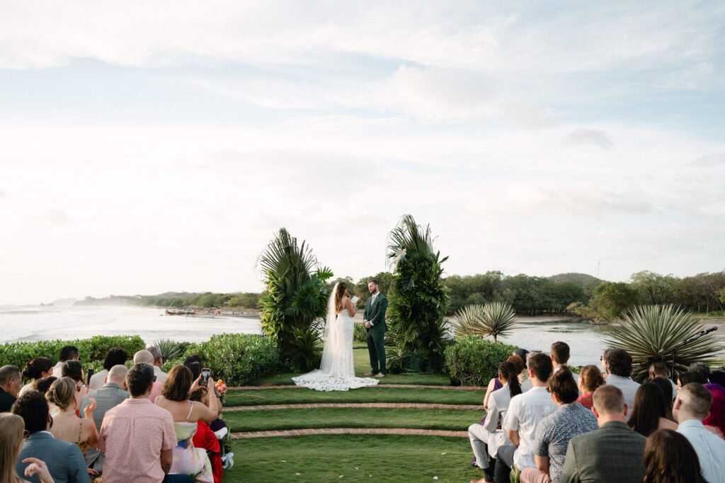 A bride and groom stand together during an outdoor wedding ceremony, surrounded by greenery and palm plants, with guests seated on either side and a scenic waterfront view in the background.