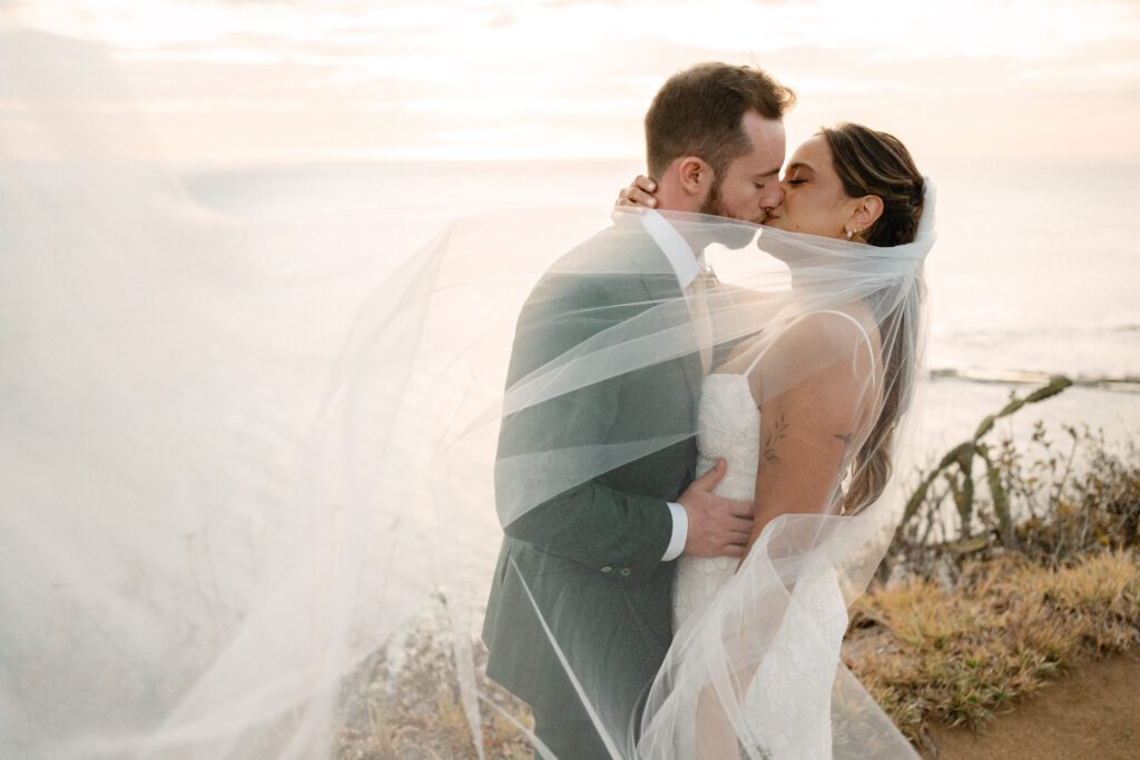 A bride and groom kiss on a cliffside at sunset, with the bride’s veil flowing around them and the ocean in the background.