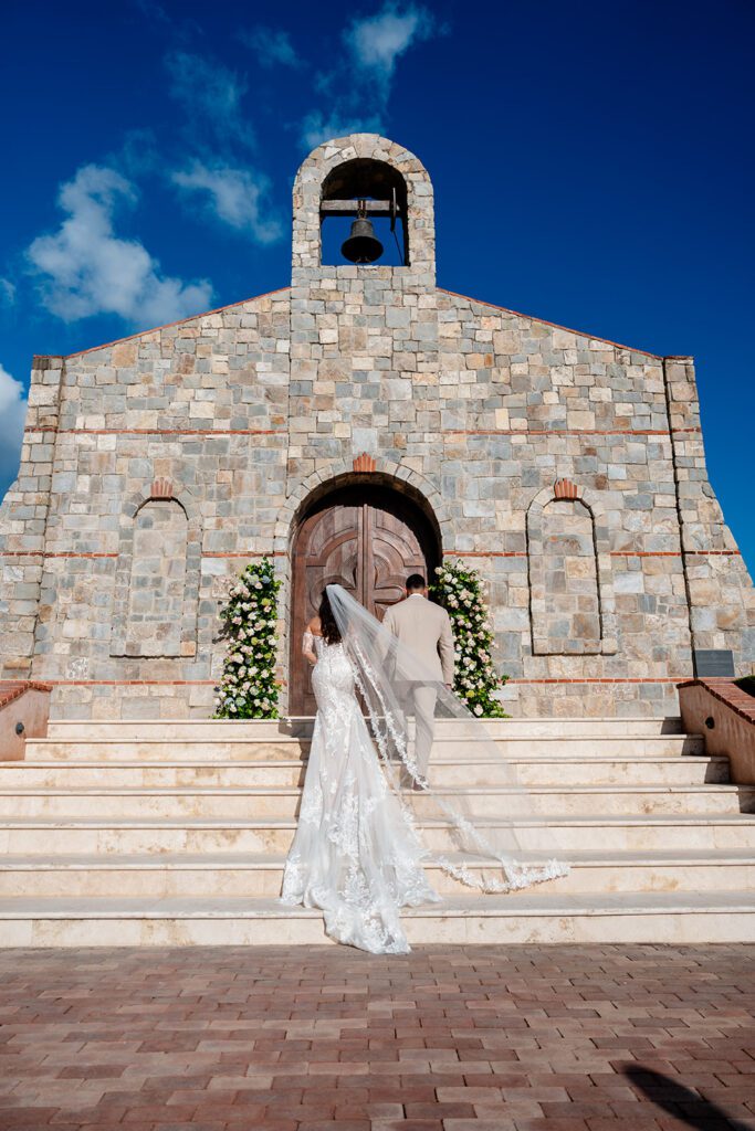 A bride and groom walk up stone steps toward a rustic stone chapel adorned with white floral arrangements, under a bright blue sky with scattered clouds.