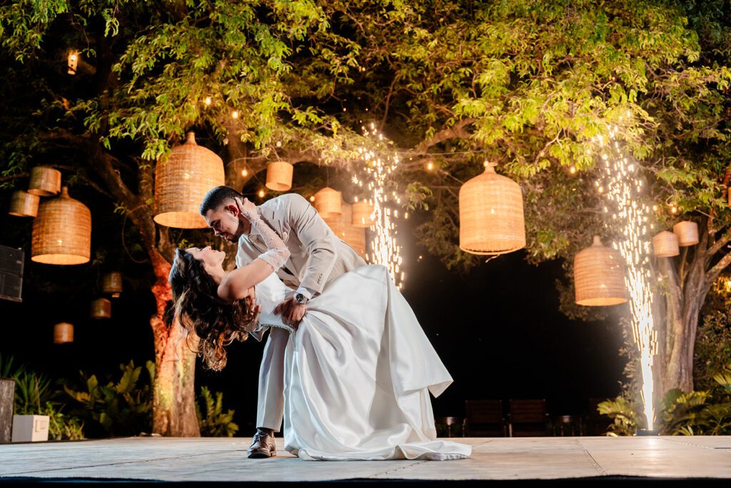 A groom dips his bride on an outdoor stage during their wedding dance, surrounded by hanging lanterns and sparkling fireworks. Trees frame the romantic moment—dream of your own and Request A Proposal today.