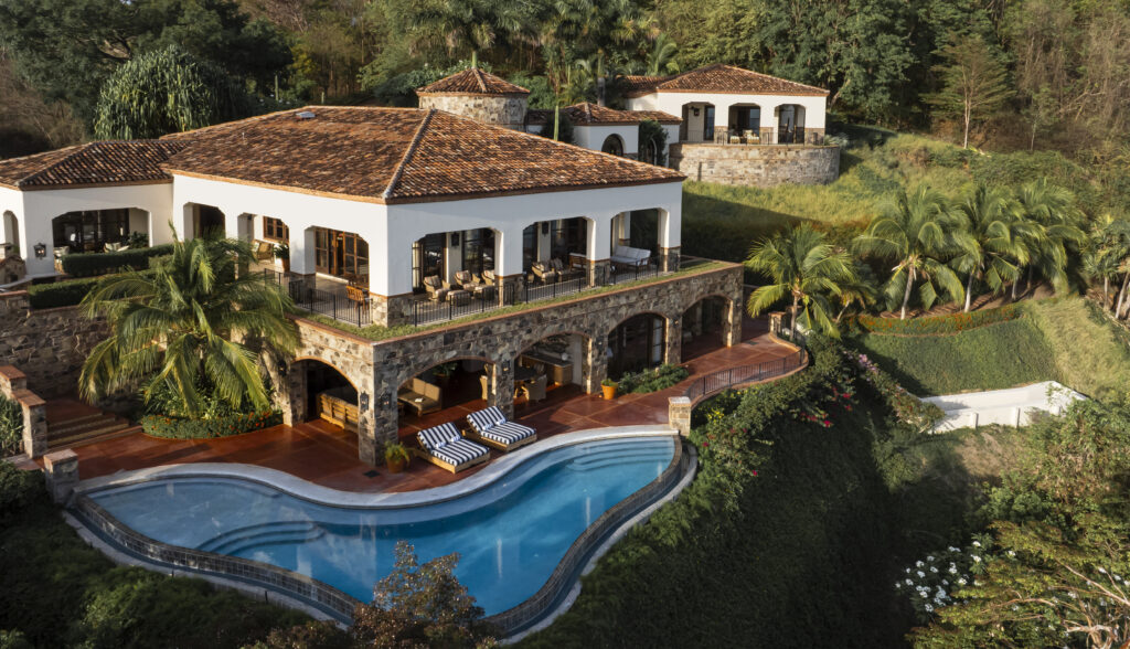 Villa Escondida is a large, luxurious co-owned luxury home with terracotta roofs and stone arches, perched on a lush hillside and featuring a curved infinity pool, sun loungers, palm trees, and vibrant green landscaping.