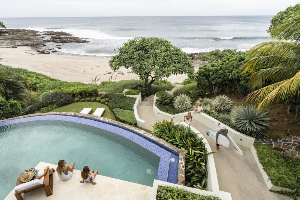 View from above of a tropical beach house: three people relax by a curved pool, while two surfers walk along a path toward the sandy beach and ocean waves.