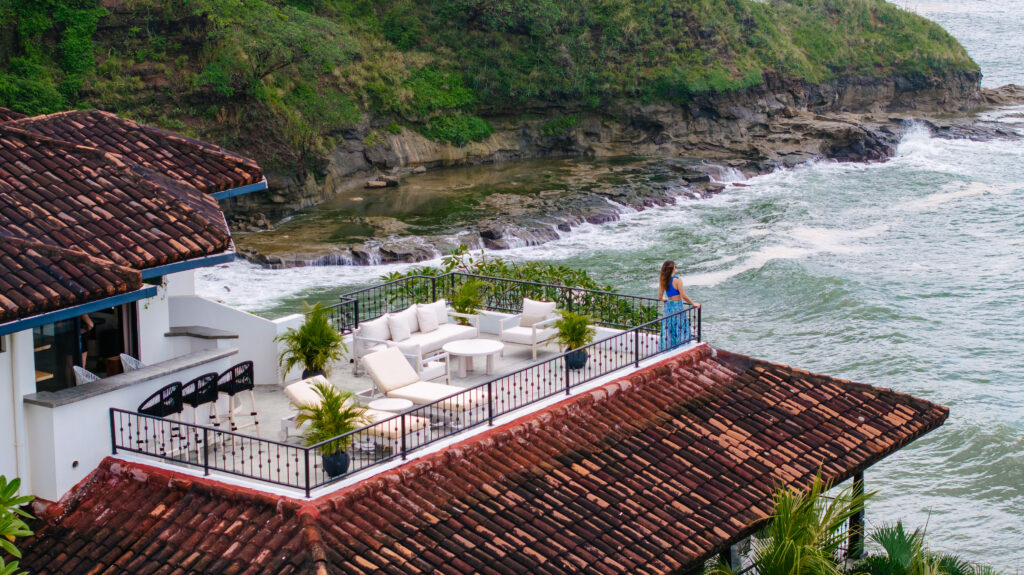 A woman stands on a spacious terrace with white outdoor furniture, overlooking the ocean and rocky shoreline of a tropical getaway, surrounded by lush greenery and red-tiled roofs.