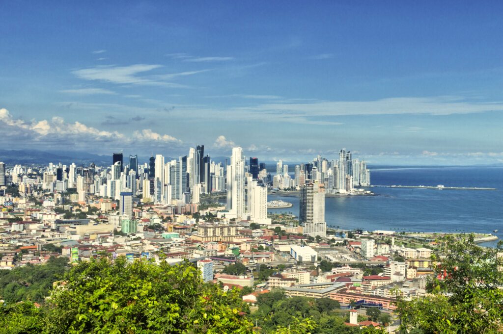 A panoramic view of Panama City, one of the best places in Central America for a vacation, features modern skyscrapers along the coastline, blue ocean waters on the right, and lush greenery in the foreground under a clear, sunny sky.