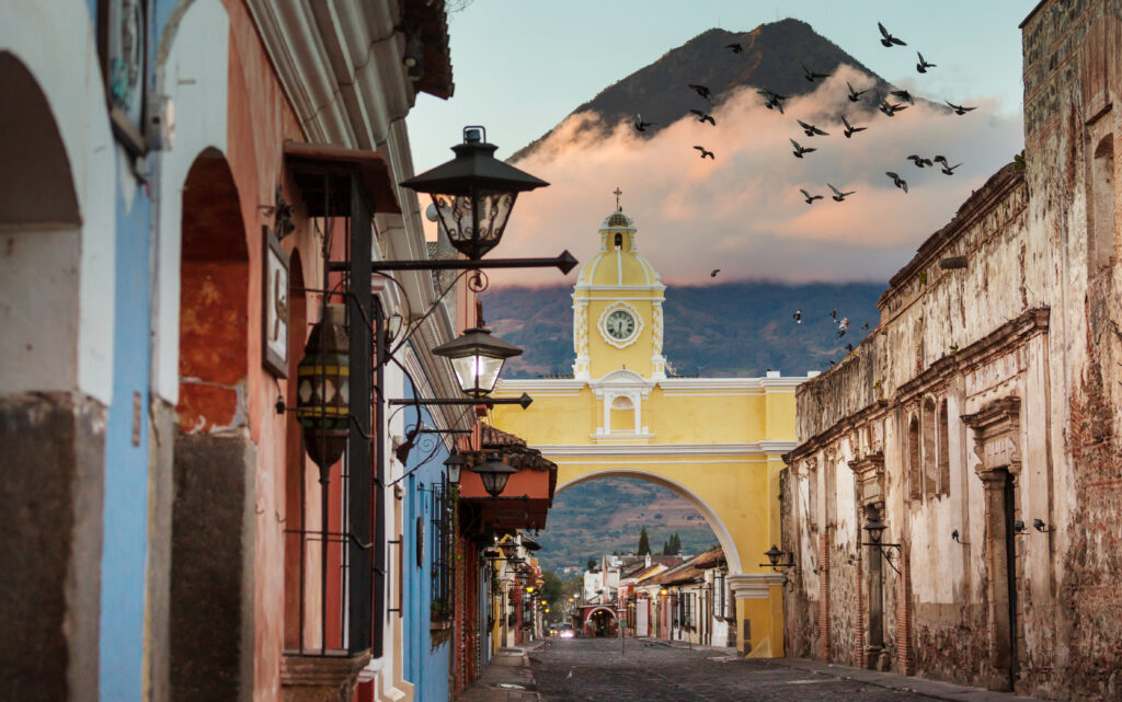 A cobblestone street in Antigua, Guatemala, features the yellow Santa Catalina Arch, with a volcano and birds flying in the background; colorful buildings line the street in one of Central Americas top destinations.