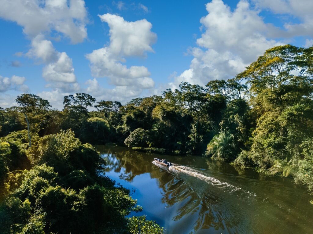 A boat travels along a river surrounded by dense green forest under a bright blue sky filled with fluffy clouds—one of the best places to experience nature’s beauty on a Central America vacation. The boat’s wake creates ripples reflecting the trees and sky.