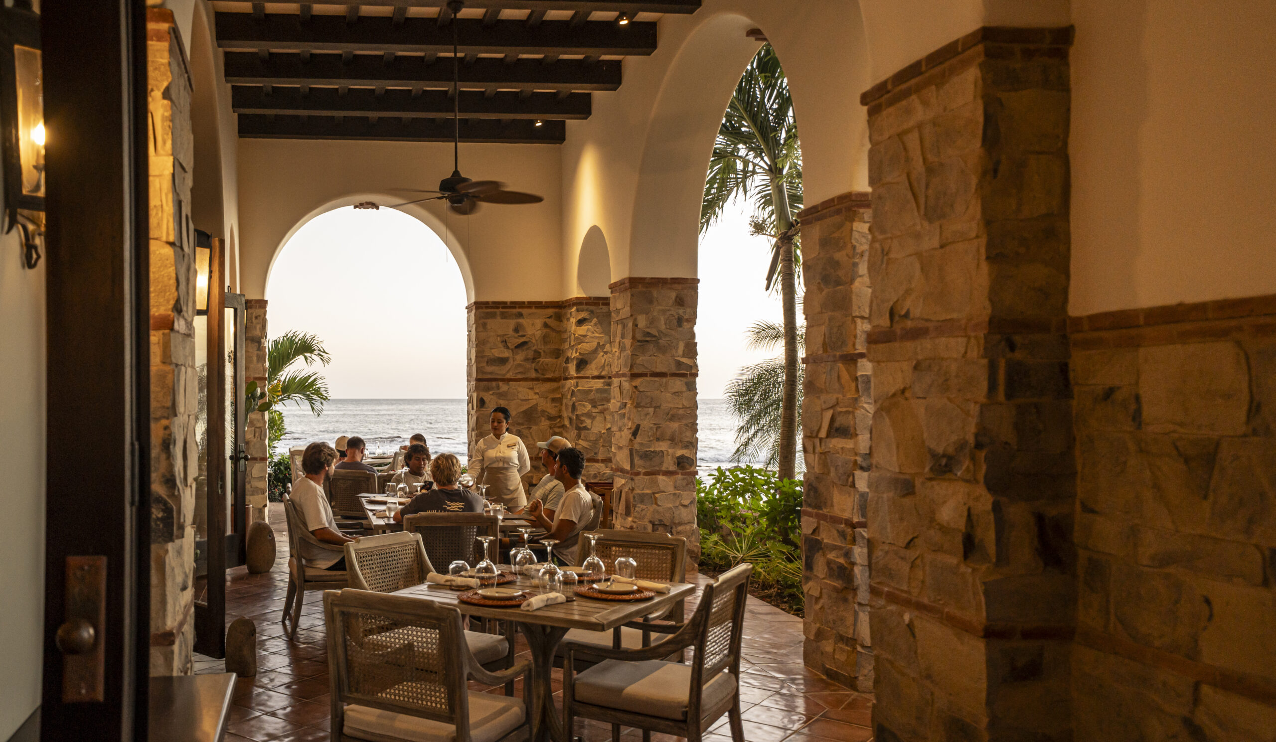 People dine at a restaurant with stone arches and columns, overlooking the ocean at sunset. Tables are set with food and drinks, and a server attends to guests. Lush greenery and palm trees are visible outside.