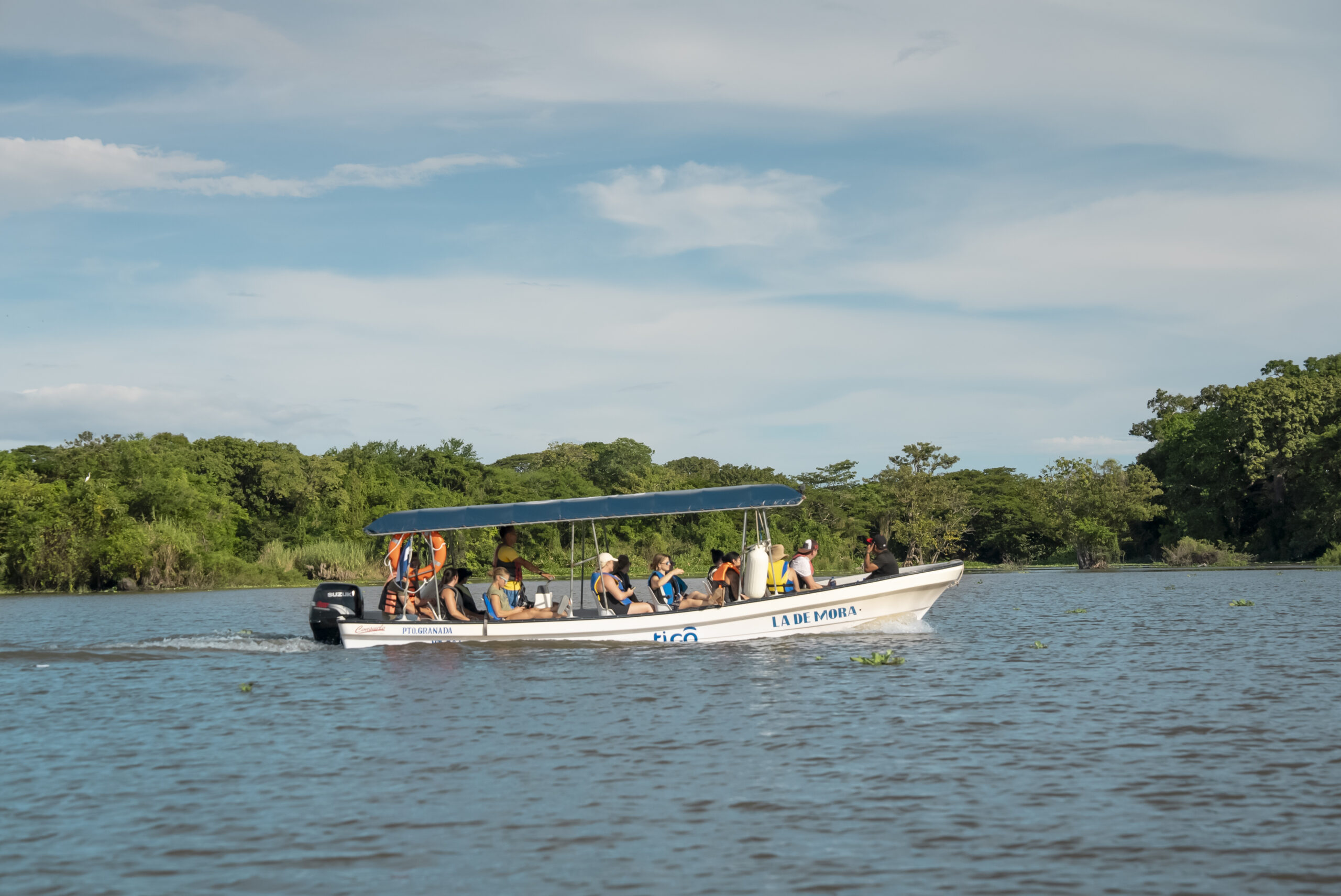 A group of people wearing life jackets ride a white boat with a blue canopy on a wide river, surrounded by lush green trees under a partly cloudy sky—perfect for memorable corporate events.