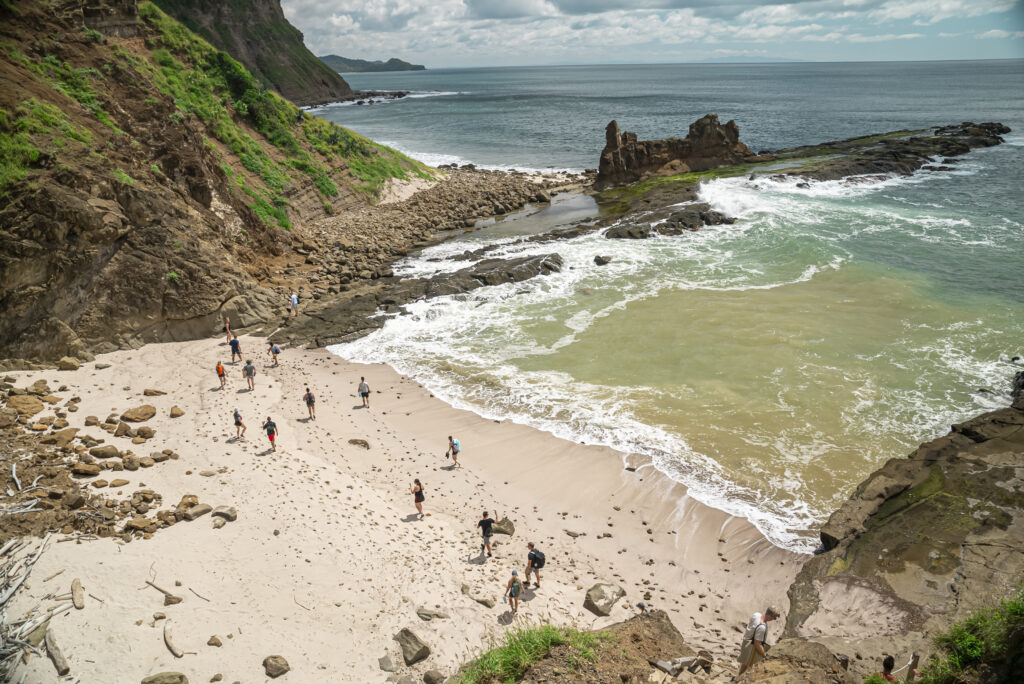 People walk along a sandy beach surrounded by rocky cliffs and green hills, with waves crashing onto the shore—a stunning backdrop often chosen for unique corporate events—while the ocean stretches into the distance under a partly cloudy sky.