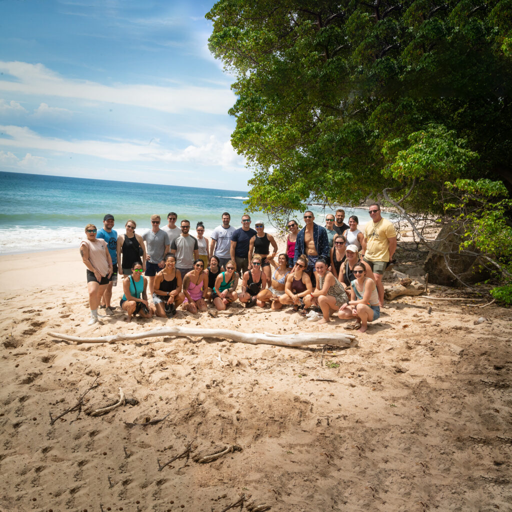 A group of people poses together on a sandy beach near the ocean during corporate events, with trees providing shade to the right and a blue sky with some clouds overhead.