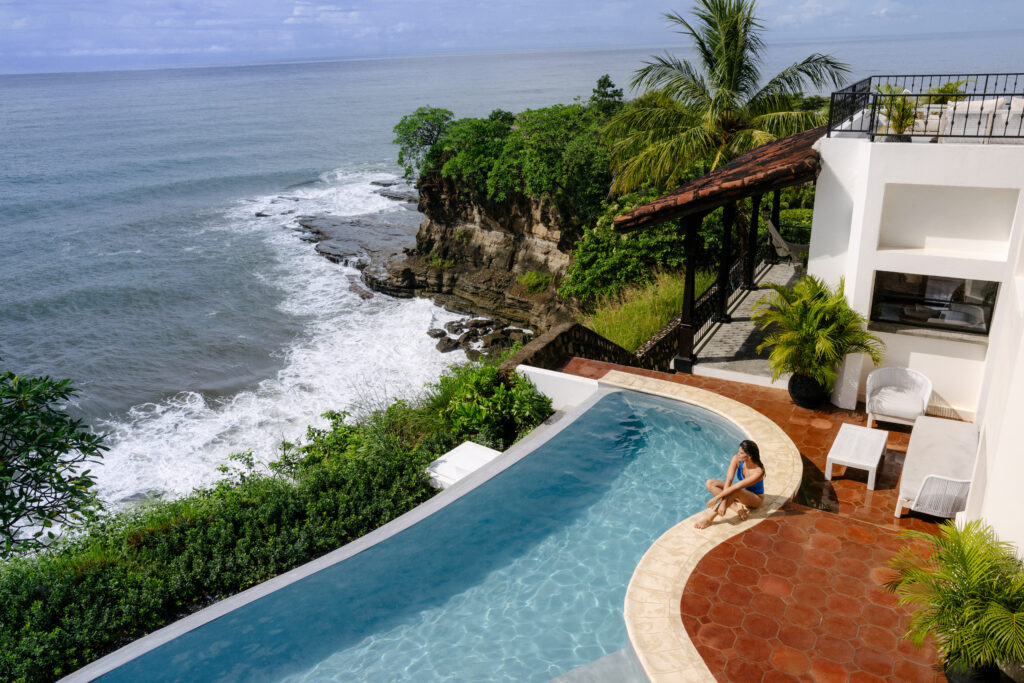 A woman sits by an infinity pool overlooking the ocean, with waves crashing against rocky cliffs and lush greenery surrounding a modern white villa.