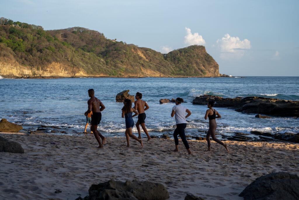 Five people jog along a sandy beach near the shoreline, embracing their wellness journey with FitSouls. Rocky cliffs and green hills rise in the background as waves crash gently on the rocks beneath the clear sky.