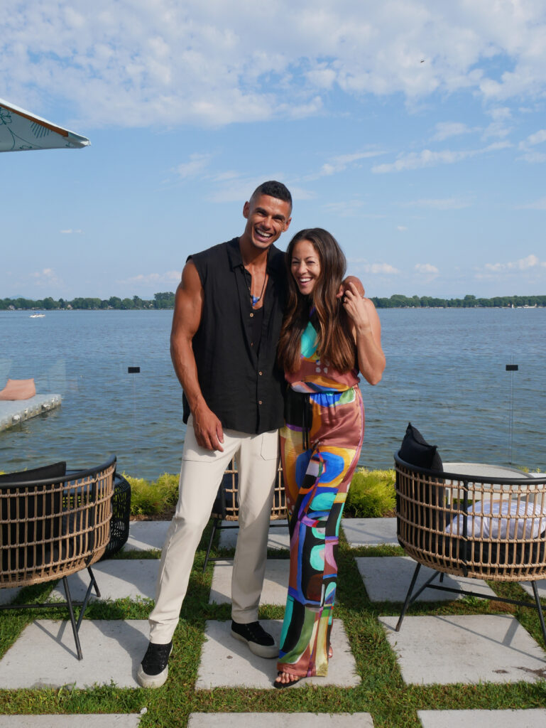 A smiling man and woman stand together outdoors by a large body of water, with modern patio chairs nearby and a partly cloudy sky above. The man wears a sleeveless black shirt and light pants; the woman wears a colorful patterned outfit.